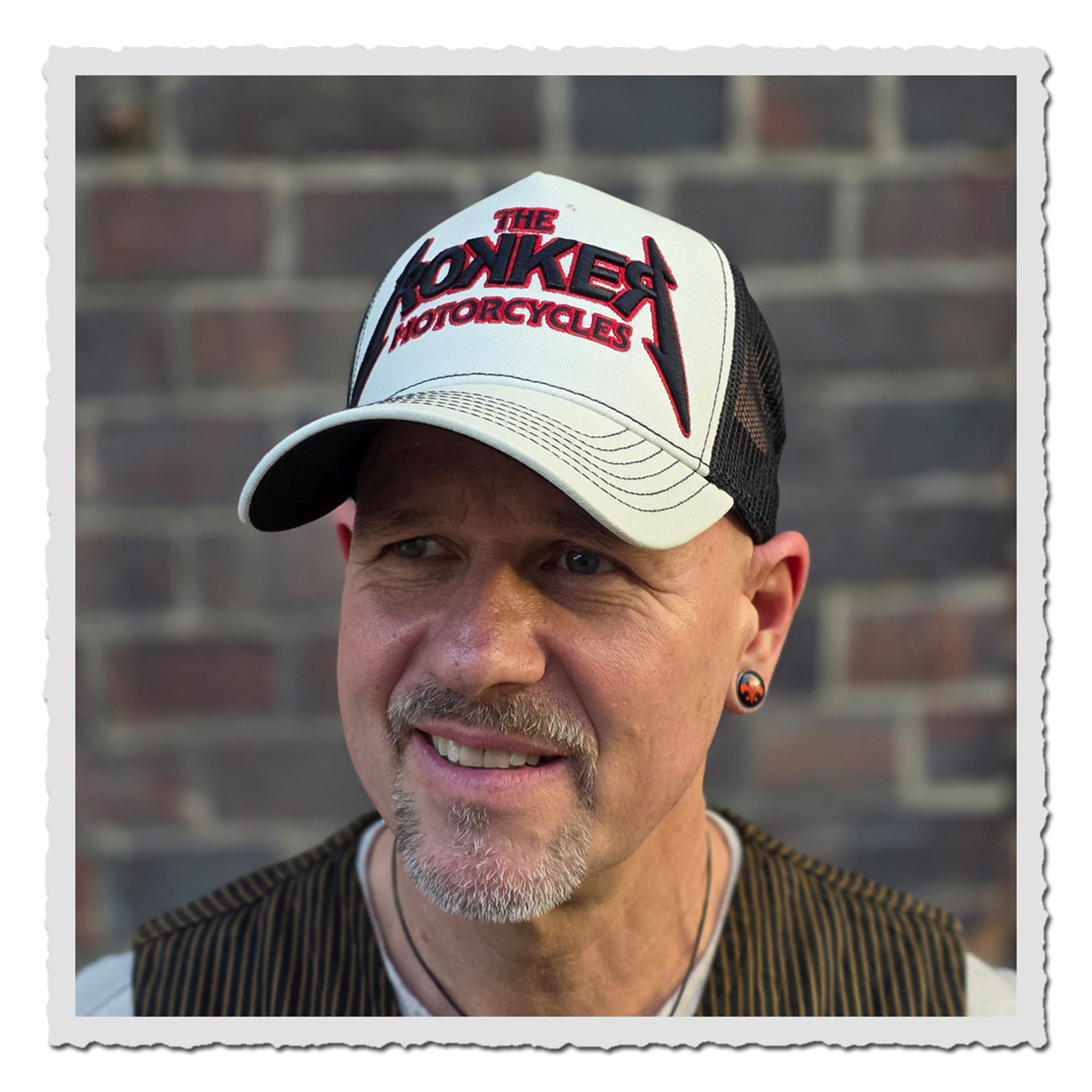 Rokker Cap Rokkallica –  Mann vor Backsteinwand trägt Retro-Trucker Cap in Grau-Schwarz 